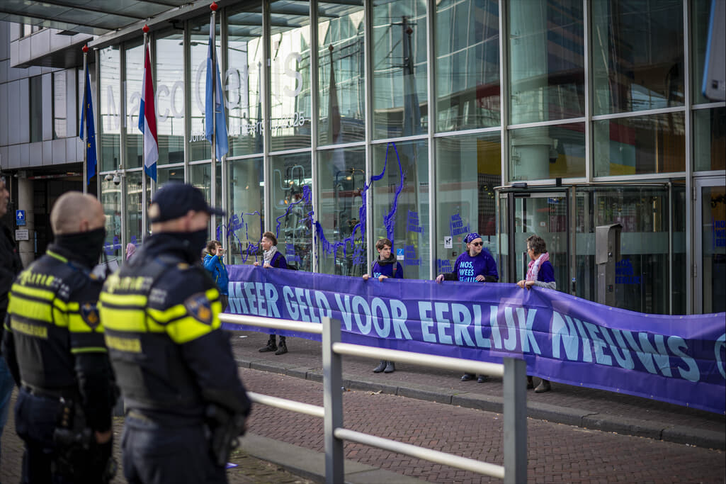 Rebellen met een banner met de tekst “Meer Geld Voor Eerlijk Nieuws” met op de voorgrond twee politieagenten op de rug gezien.