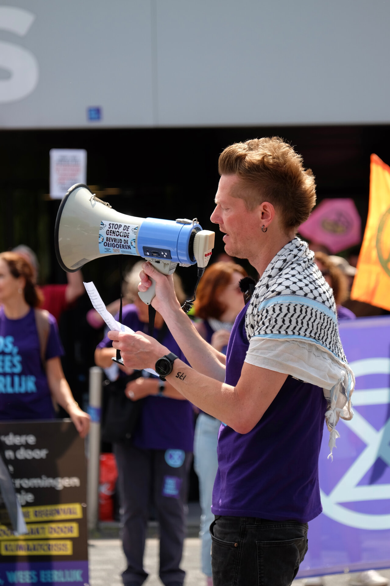 Martijn Dekker met megafoon in zijn rechterhand en A4 papier met tekst in zijn linkerhand. Hij draagt een paars t-shirt en een keffiyeh over zijn schouders. In de achtergrond zijn rebellen te zien.