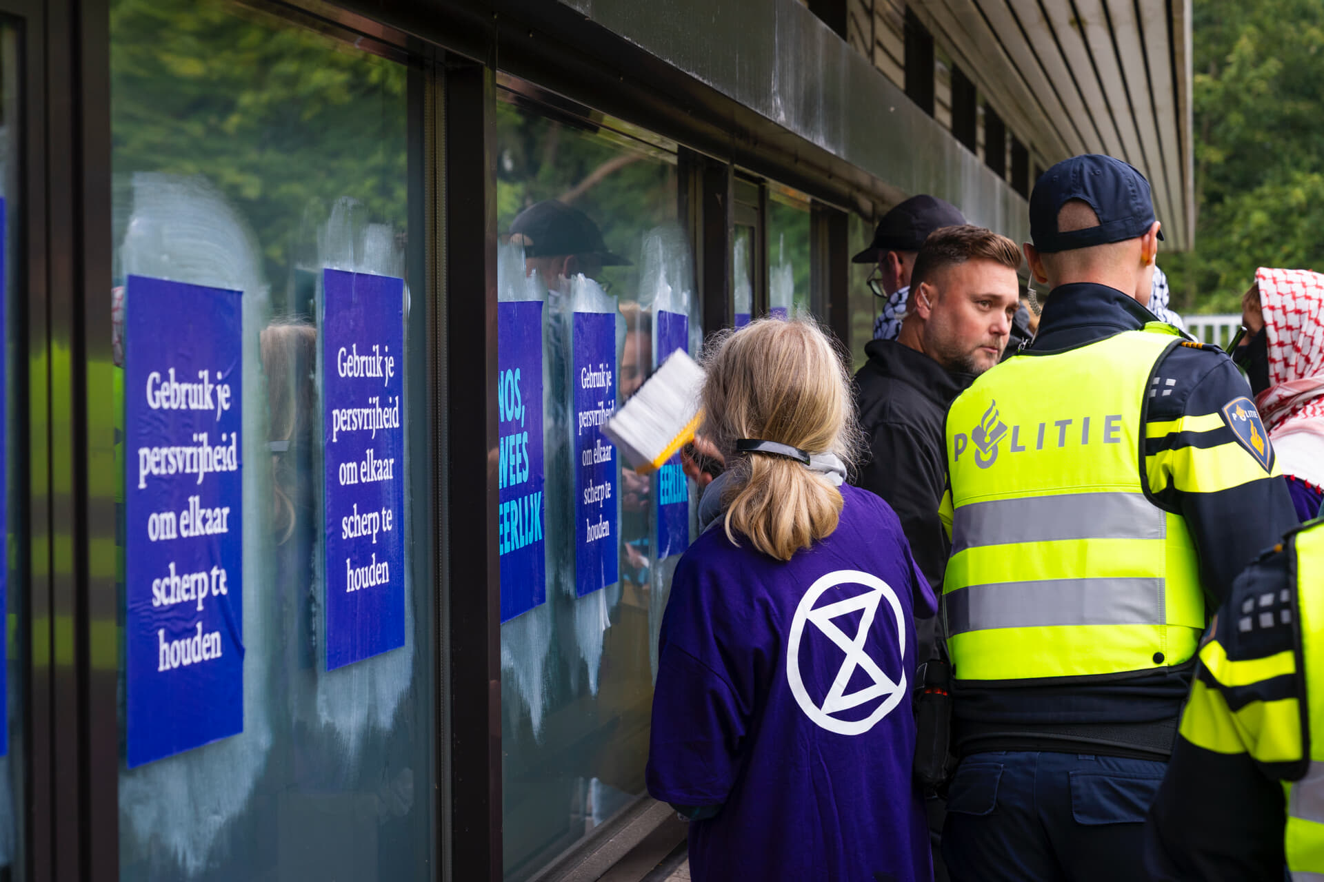 Rebel plakt posters met de tekst “Gebruik je persvrijheid om elkaar scherp te houden” en “NOS, Weer Eerlijk” op de ramen van het NOS geebouw met een grote kwast met behangsellijm. Rechts in beeld is een politieagent op de rug te zien met daarachter andere rebellen.